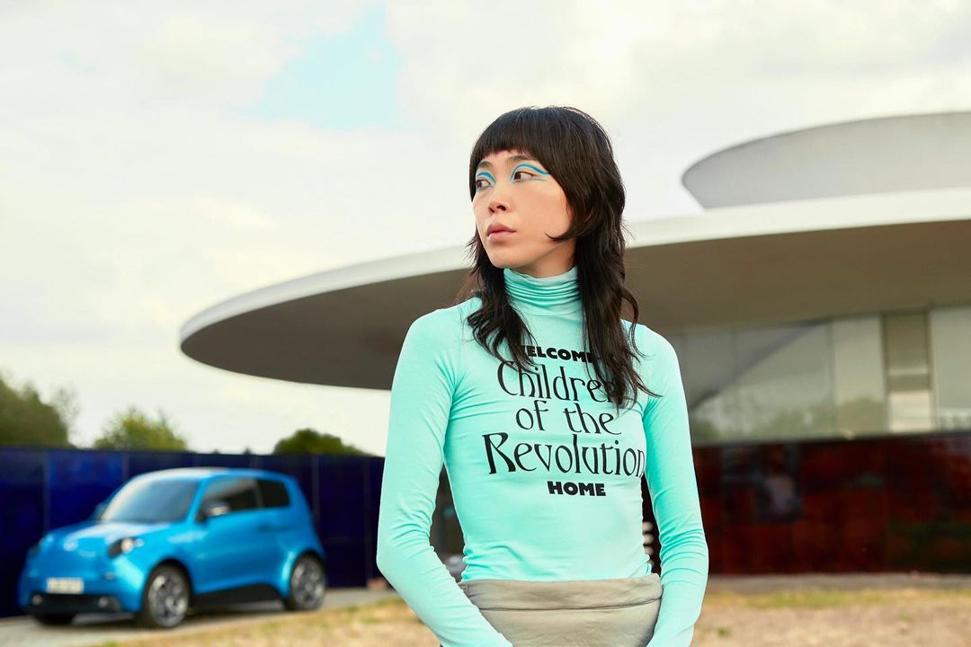 A woman in a blue top stands in front of a modern building and a blue electric car, set against a bright sky with scattered clouds.