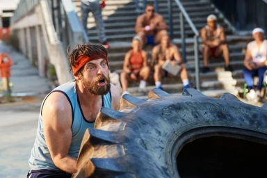 Bearded man in light blue tank top and red headband straining while flipping a large tire outdoors