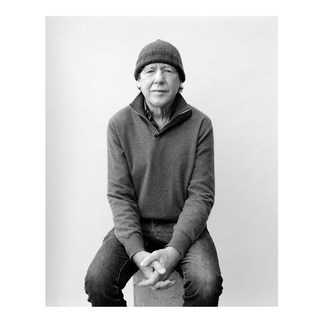 Portrait of an elderly man wearing a beanie and zip-up sweater seated against a plain background by Jody Rogac.