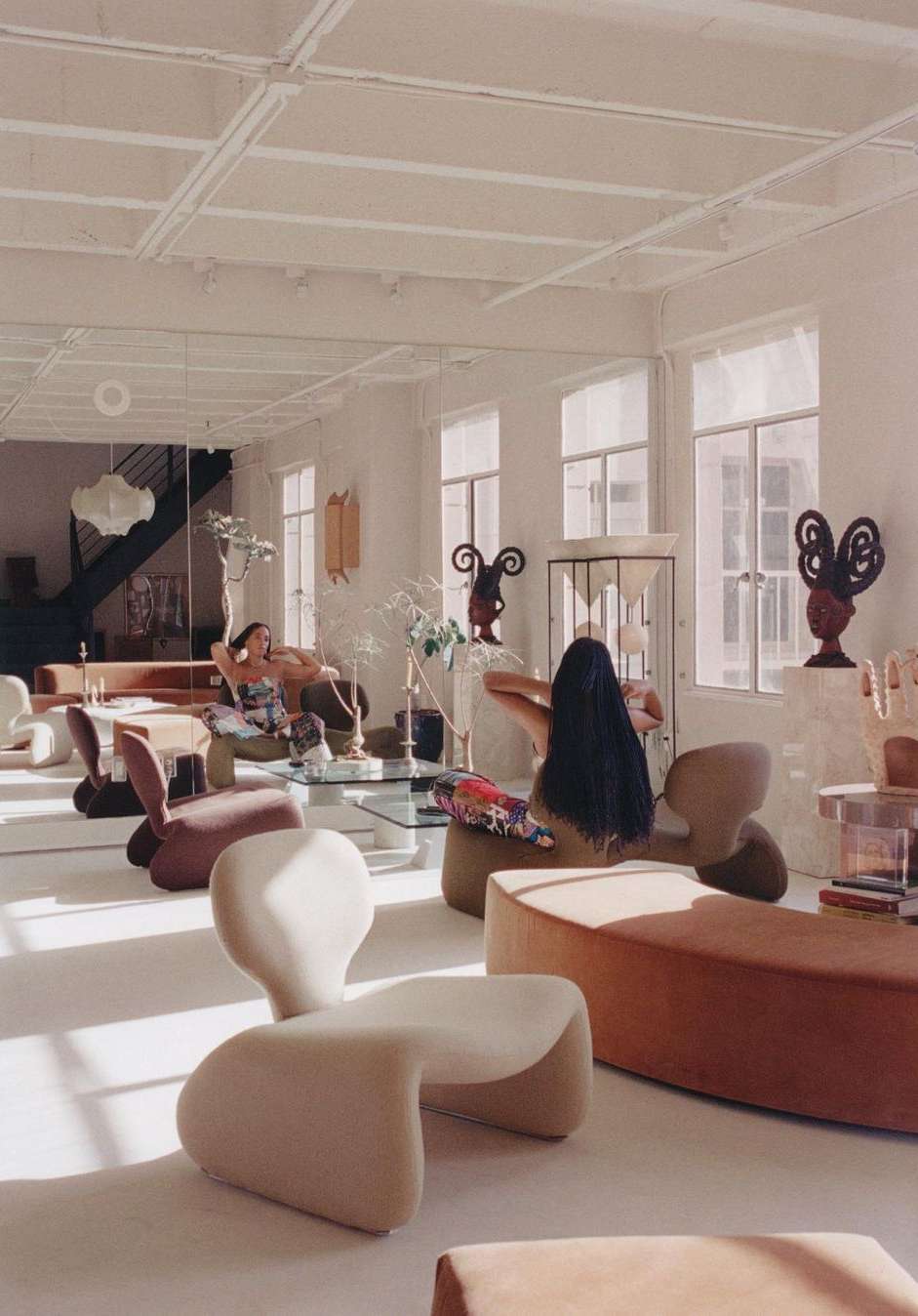 Two women relaxing in a bright sunlit studio apartment filled with sculptural furniture and decorative objects, creating a modern, airy living space.
