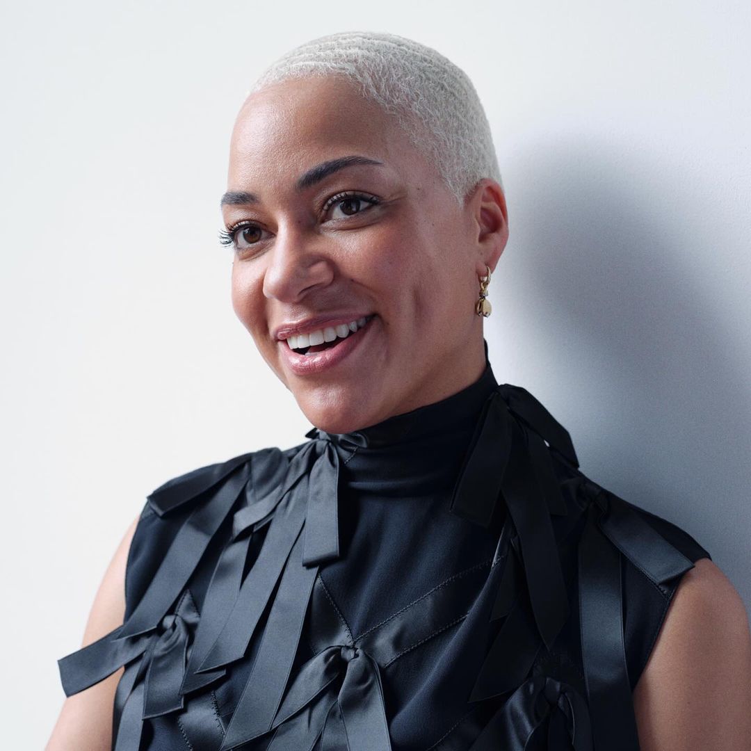 Portrait of a smiling woman with short silver hair wearing a black satin sleeveless top against a white wall.