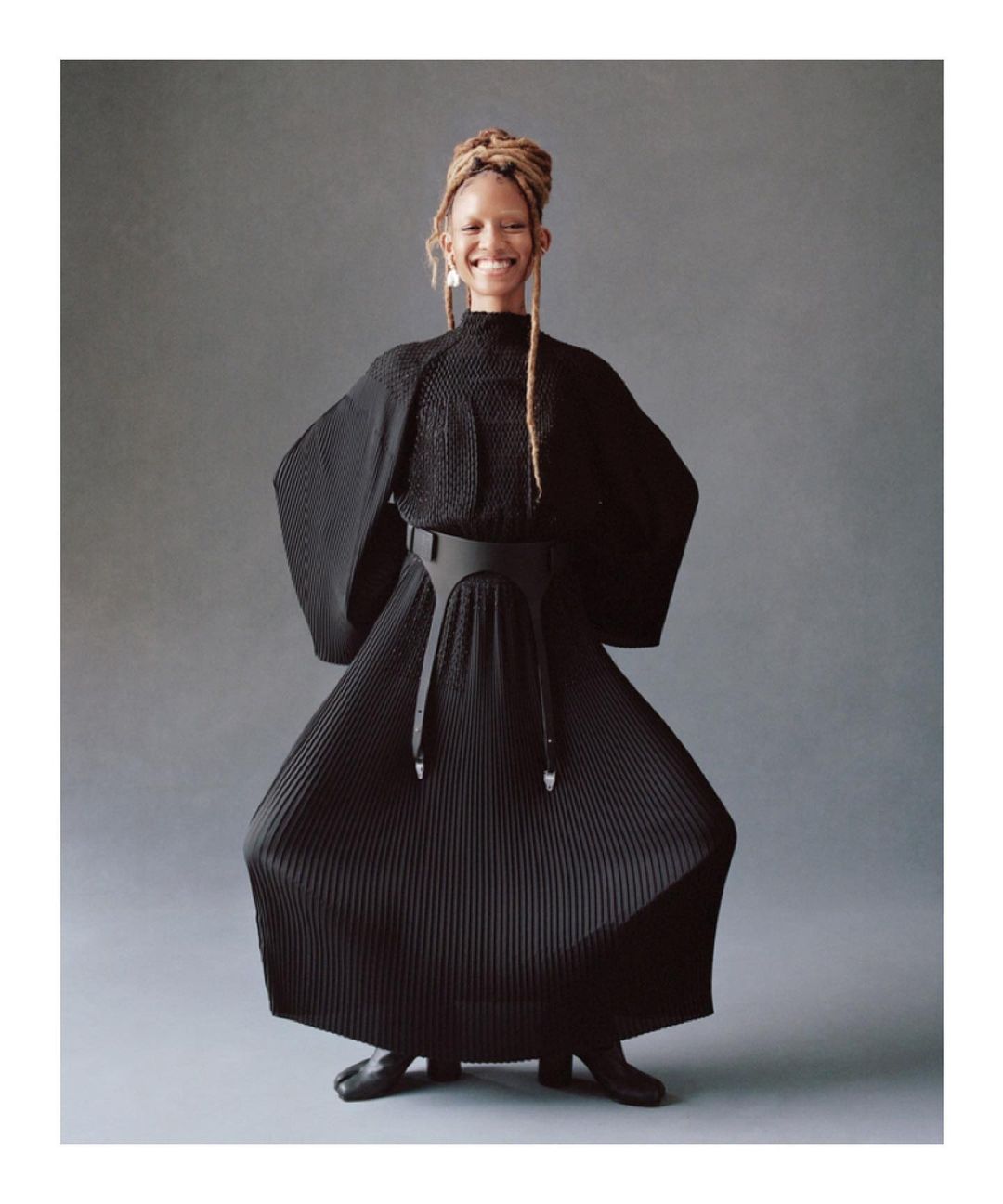 Woman in black dress and leather harness belt poses with a smile against gray background in a full shot. Studio lighting.