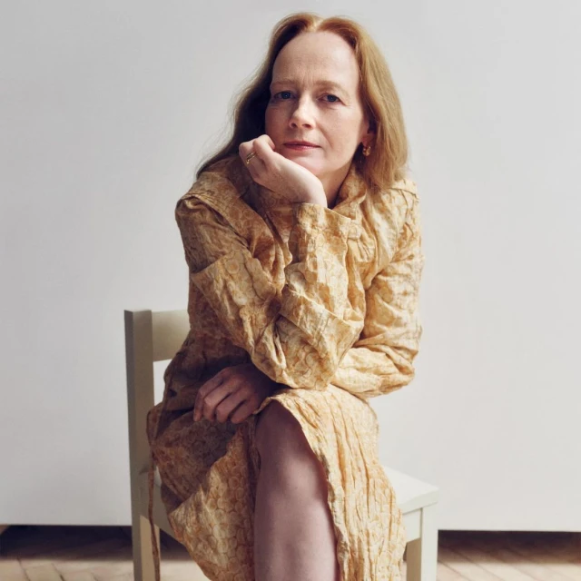 Portrait of a middle-aged woman with auburn hair, wearing a textured yellow dress, sitting on a chair against a white background in a photo studio.