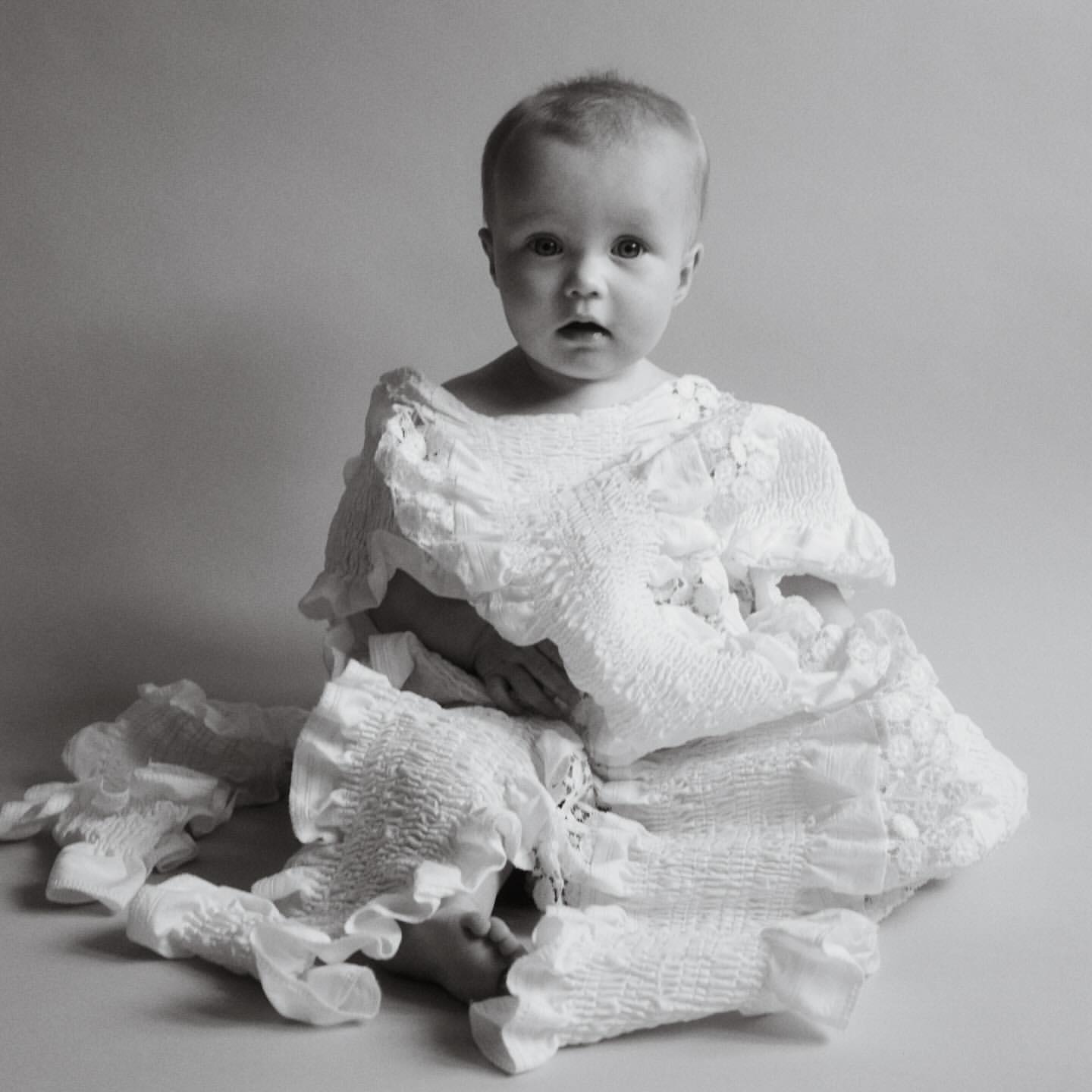 Black and white studio portrait of a baby wearing a white ruffled garment, looking directly at the camera with a neutral expression.
