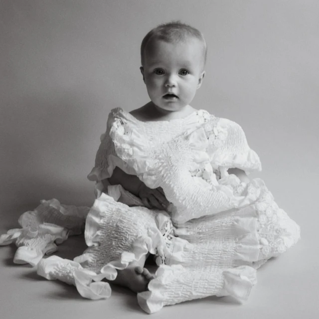 Black and white studio portrait of a baby wearing a white ruffled garment, looking directly at the camera with a neutral expression.