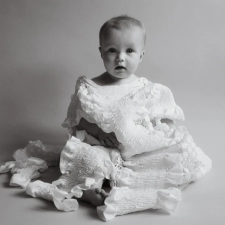 Black and white studio portrait of a baby wearing a white ruffled garment, looking directly at the camera with a neutral expression.