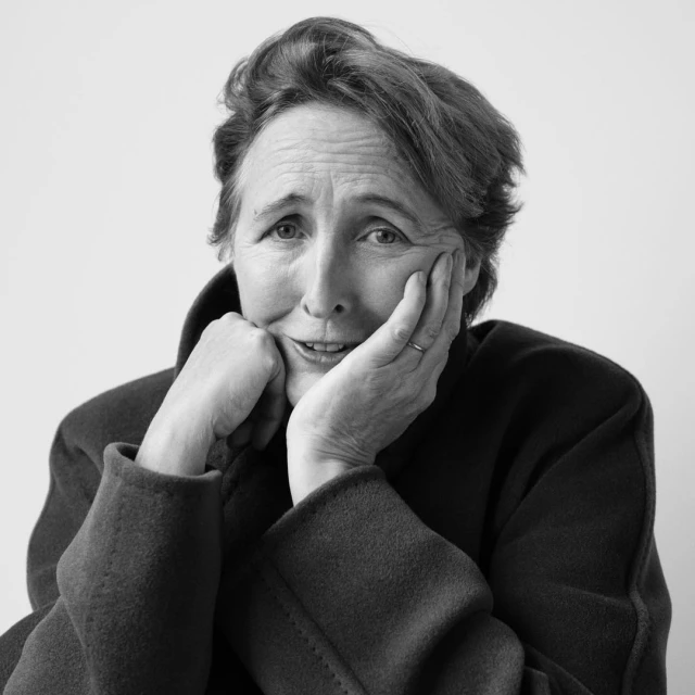 Black and white portrait of an elderly woman with short hair, resting her face on her hands, wearing a dark coat against a white background.