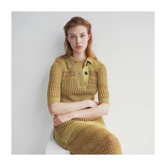 Portrait of a red-haired woman wearing a yellow-gold knit midi dress with buttons, posing indoors against a white wall in natural light.