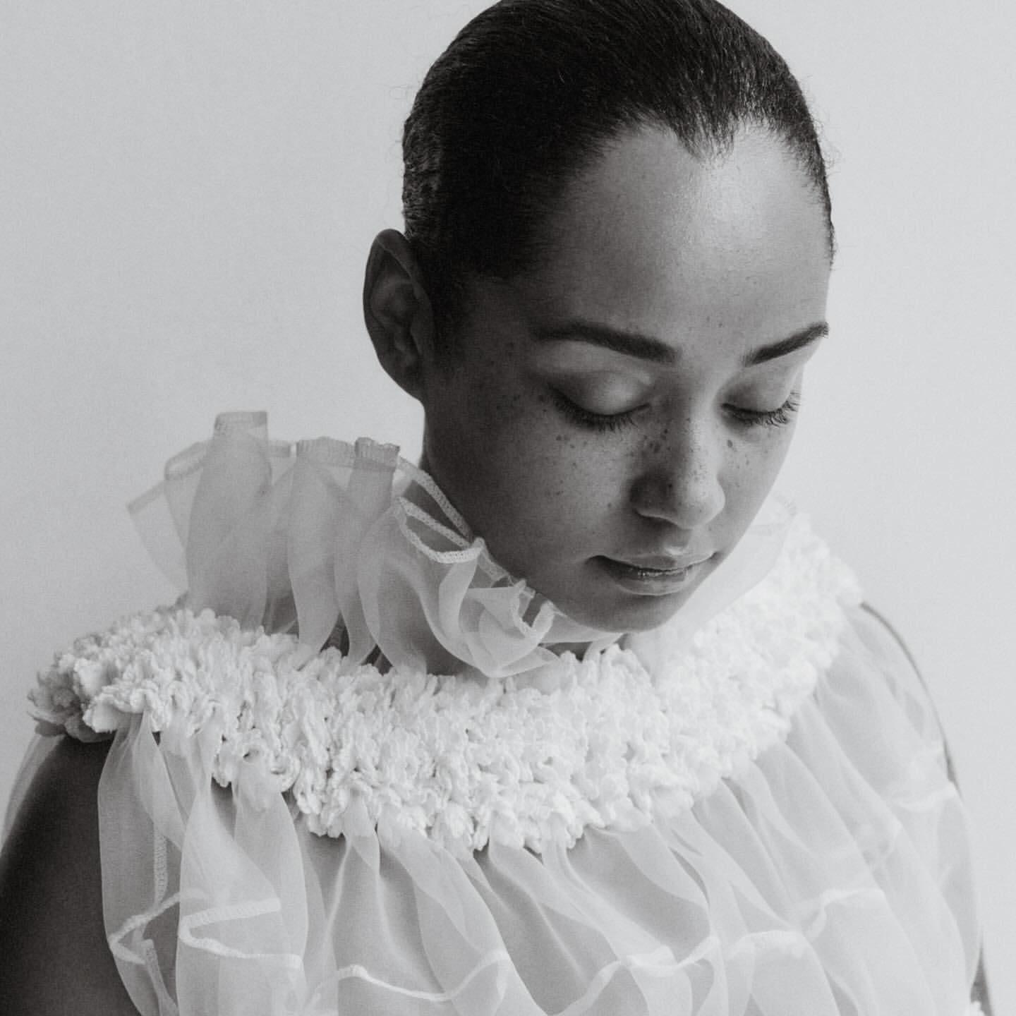 A monochrome portrait of a person with short hair, looking down, wearing a white, layered, and ruffled collar and top against a plain background.