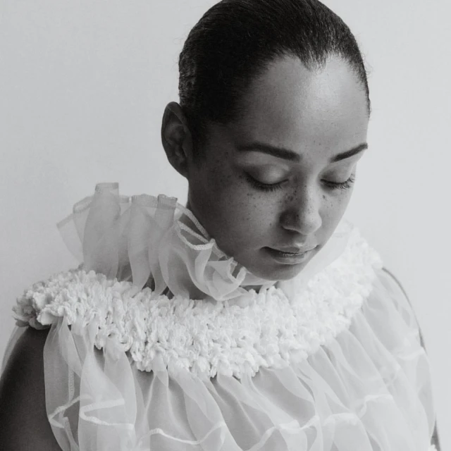 A monochrome portrait of a person with short hair, looking down, wearing a white, layered, and ruffled collar and top against a plain background.