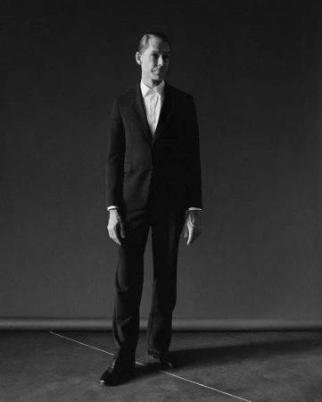 A man in a black suit and white shirt standing full-length against a dark seamless studio backdrop in New York.