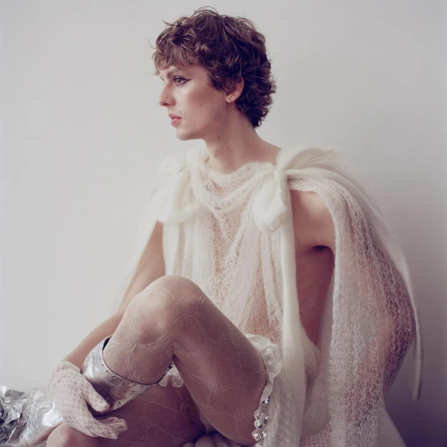 Person with curly brown hair poses in a sheer white cape and patterned hosiery against a pale background in a studio setting.
