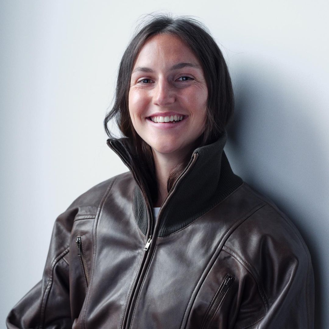 Smiling woman wearing a brown leather bomber jacket stands indoors against a white wall, captured in a medium shot.