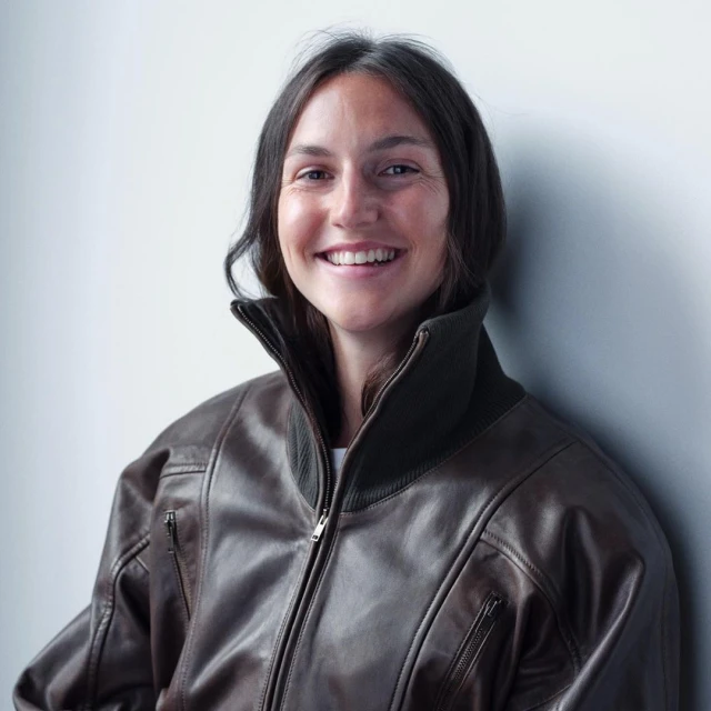 Smiling woman wearing a brown leather bomber jacket stands indoors against a white wall, captured in a medium shot.