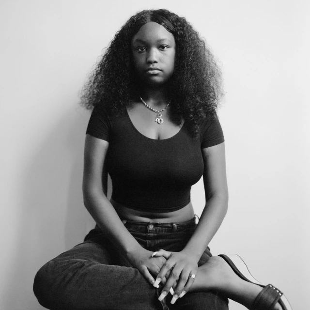Portrait of a young Black woman with curly hair in jeans and a crop top, sitting on a stool in front of a white wall, looking at the camera.