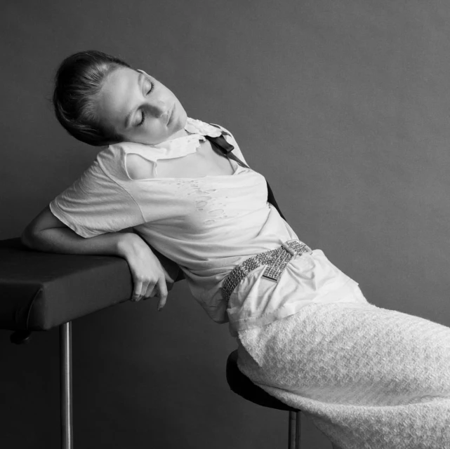 Monochrome portrait of an adult woman posing in a light-colored top and skirt. She is reclining on a padded surface indoors.