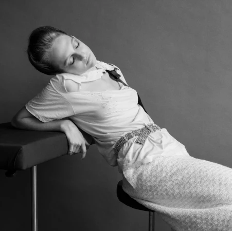 Monochrome portrait of an adult woman posing in a light-colored top and skirt. She is reclining on a padded surface indoors.