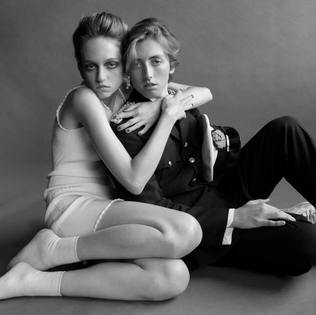 Monochrome portrait of two fair-skinned young adults, one embracing the other, seated against a grey backdrop in a studio setting.