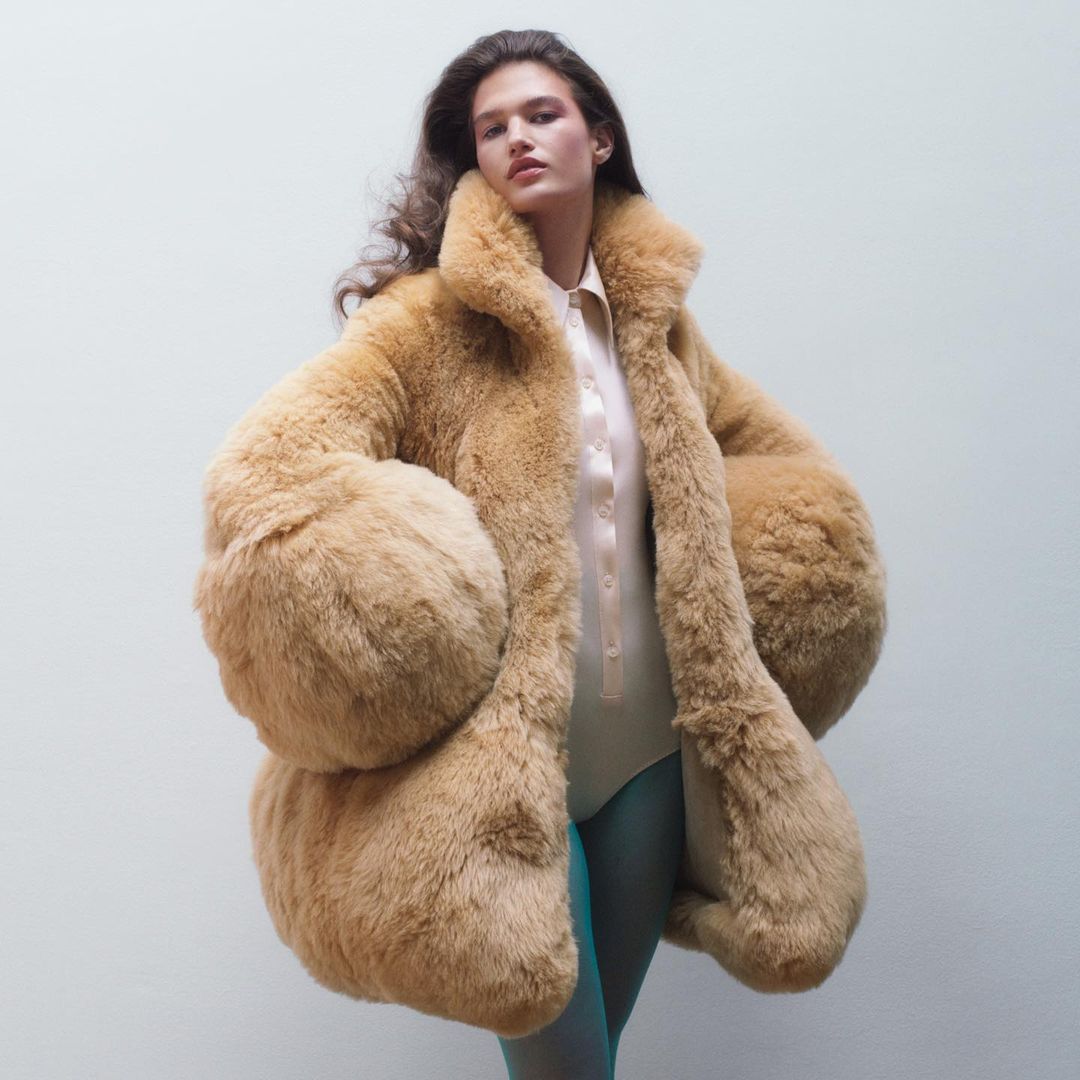 Woman in a tan faux fur coat with puffy sleeves, a pink shirt and teal tights, posing in front of a neutral studio background.