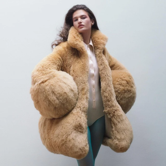 Woman in a tan faux fur coat with puffy sleeves, a pink shirt and teal tights, posing in front of a neutral studio background.