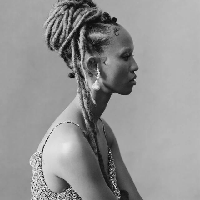 Portrait of a Black woman with dreadlocks in an updo, wearing pearl earrings and a beaded top against a grey backdrop, captured in black and white.