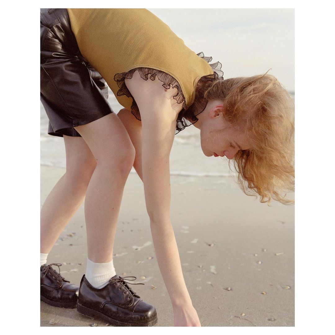 Young adult female on beach bending over in a tan ribbed ruffle shirt, black shorts, white socks, and black shoes with ocean backdrop.