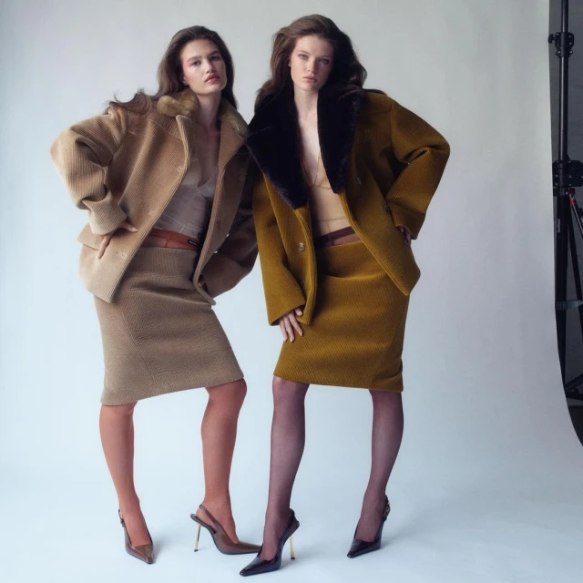 Two fashion models pose in matching skirt suits with fur-trimmed coats in a studio. One suit is tan, the other is mustard-colored.