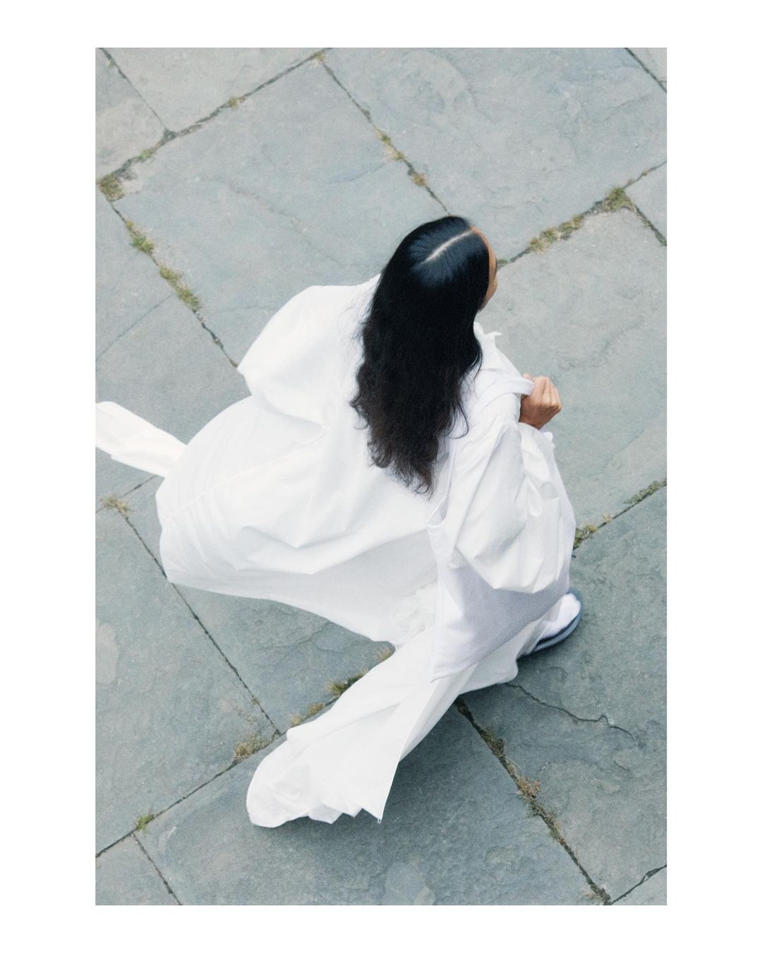 Overhead shot of a woman in a white dress crouching on gray paving stone with black hair.