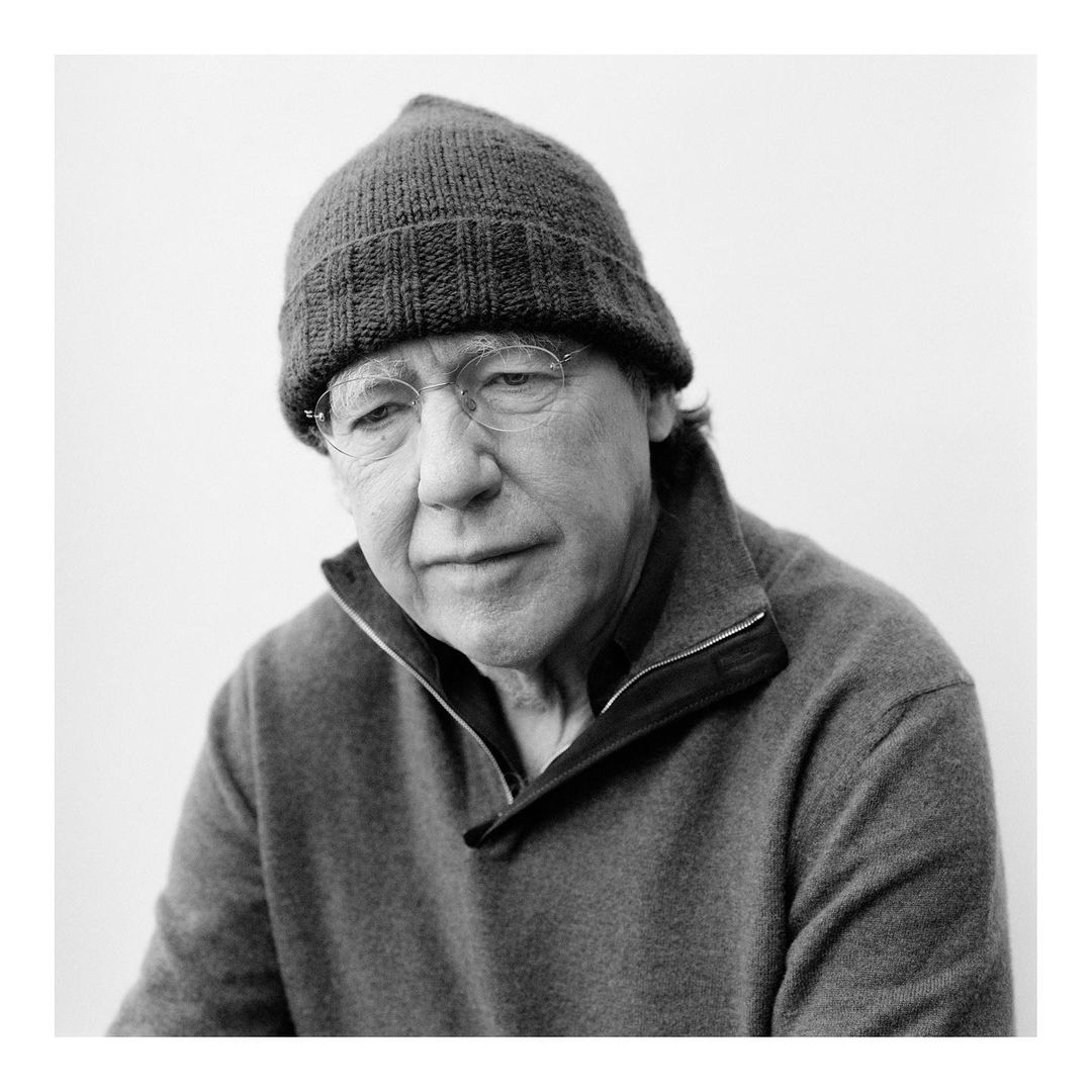 Monochrome portrait of a mature adult man with glasses wearing a knitted hat and a gray sweater against a neutral backdrop.