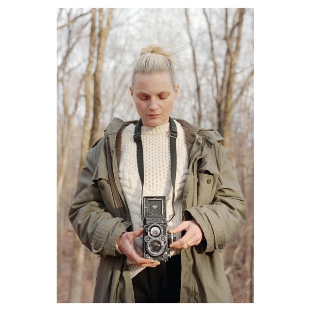 A woman stands in a forest holding a vintage twin-lens reflex camera. The woman has fair skin and blonde hair and is wearing a green jacket.