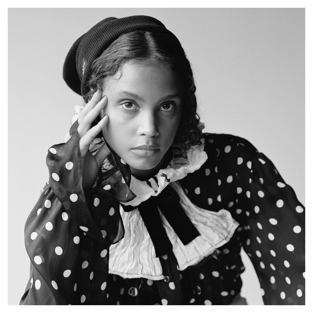Monochrome portrait of a young woman with curly hair wearing a knit hat and polka dot blouse against a bright, neutral backdrop.