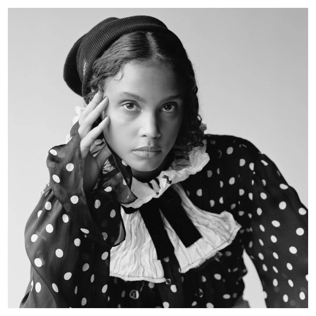 Monochrome portrait of a young woman with curly hair wearing a knit hat and polka dot blouse against a bright, neutral backdrop.