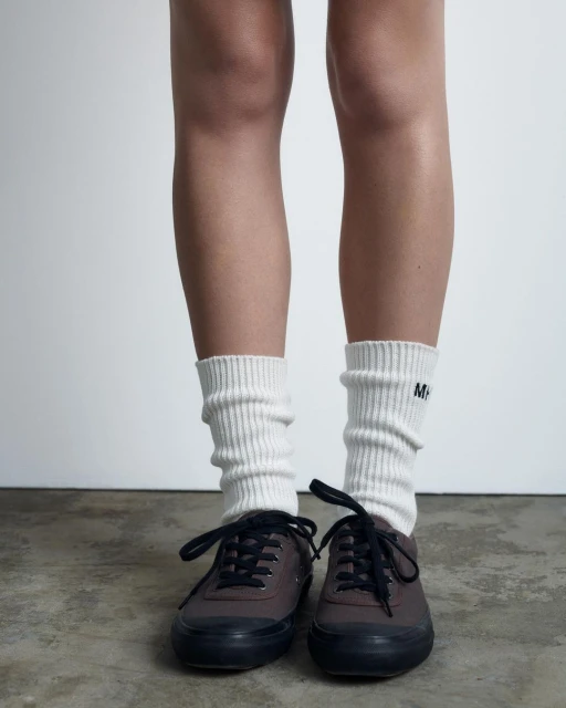 Woman's legs and feet in white ribbed socks and dark brown sneakers on a gray background, showcasing a casual, comfortable fashion choice.