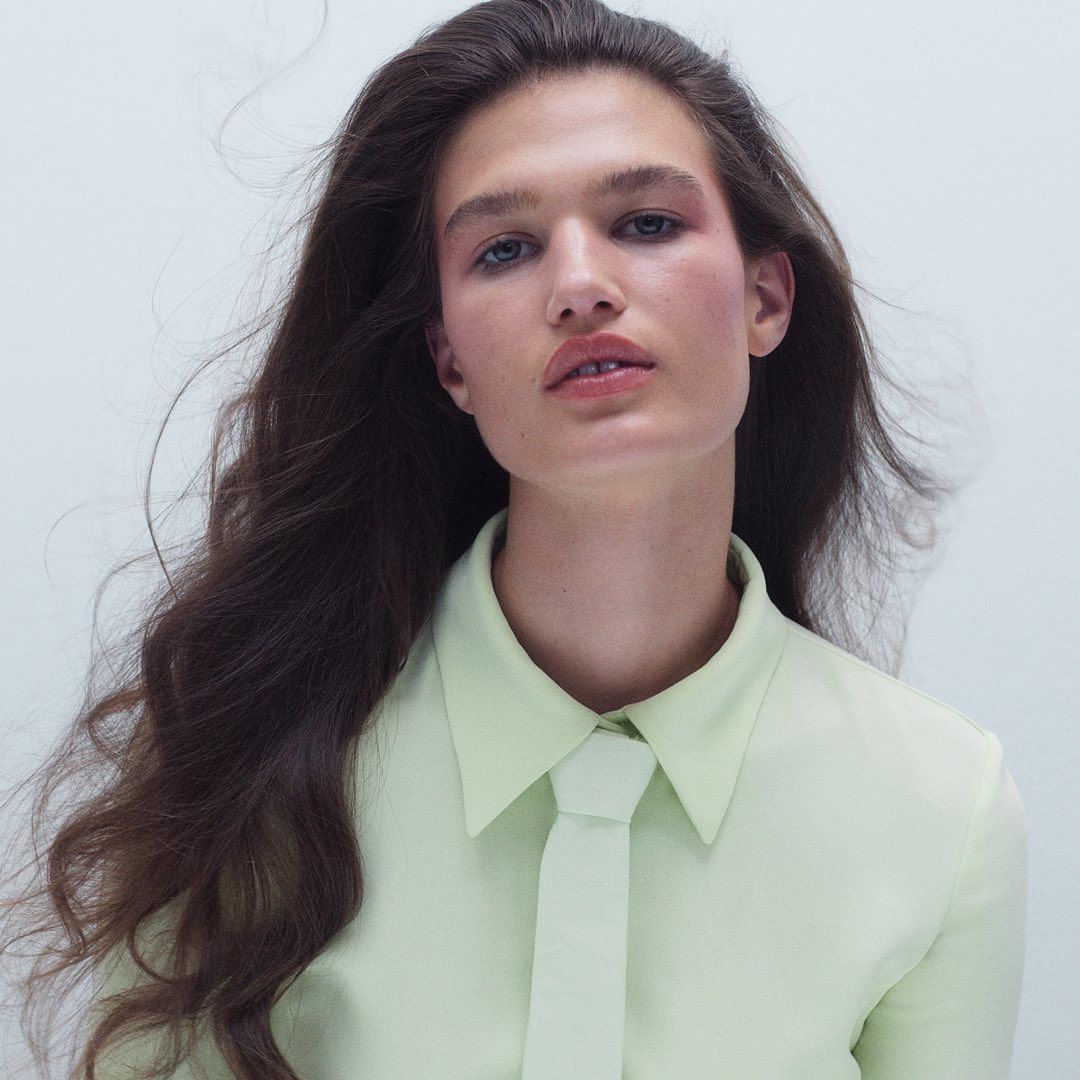 Close-up shot of a brunette adult female posing with fair skin, long wavy hair, light eye makeup, and a light green collared shirt with necktie.