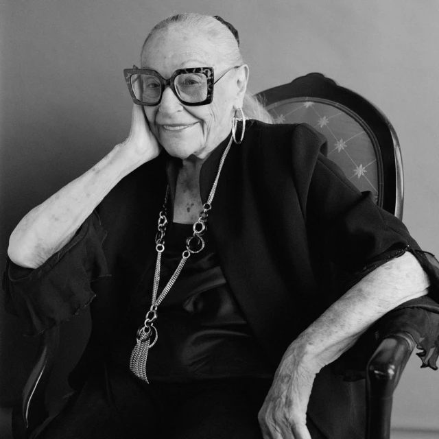 Monochrome portrait of an elderly woman with large eyeglasses seated in a chair, wearing a black jacket and statement necklace.