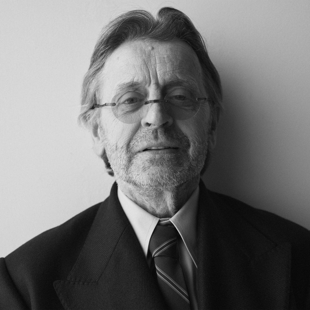 Black and white portrait of an older man with glasses wearing a suit and tie, captured at eye-level against a plain white background.