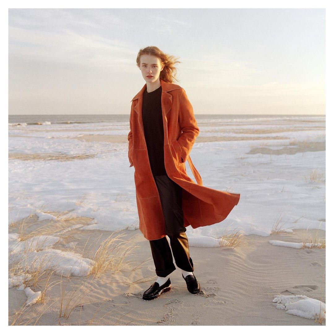 Woman in a long orange coat poses on a snow-covered beach with a cloudy sky backdrop, wearing black trousers and loafers.