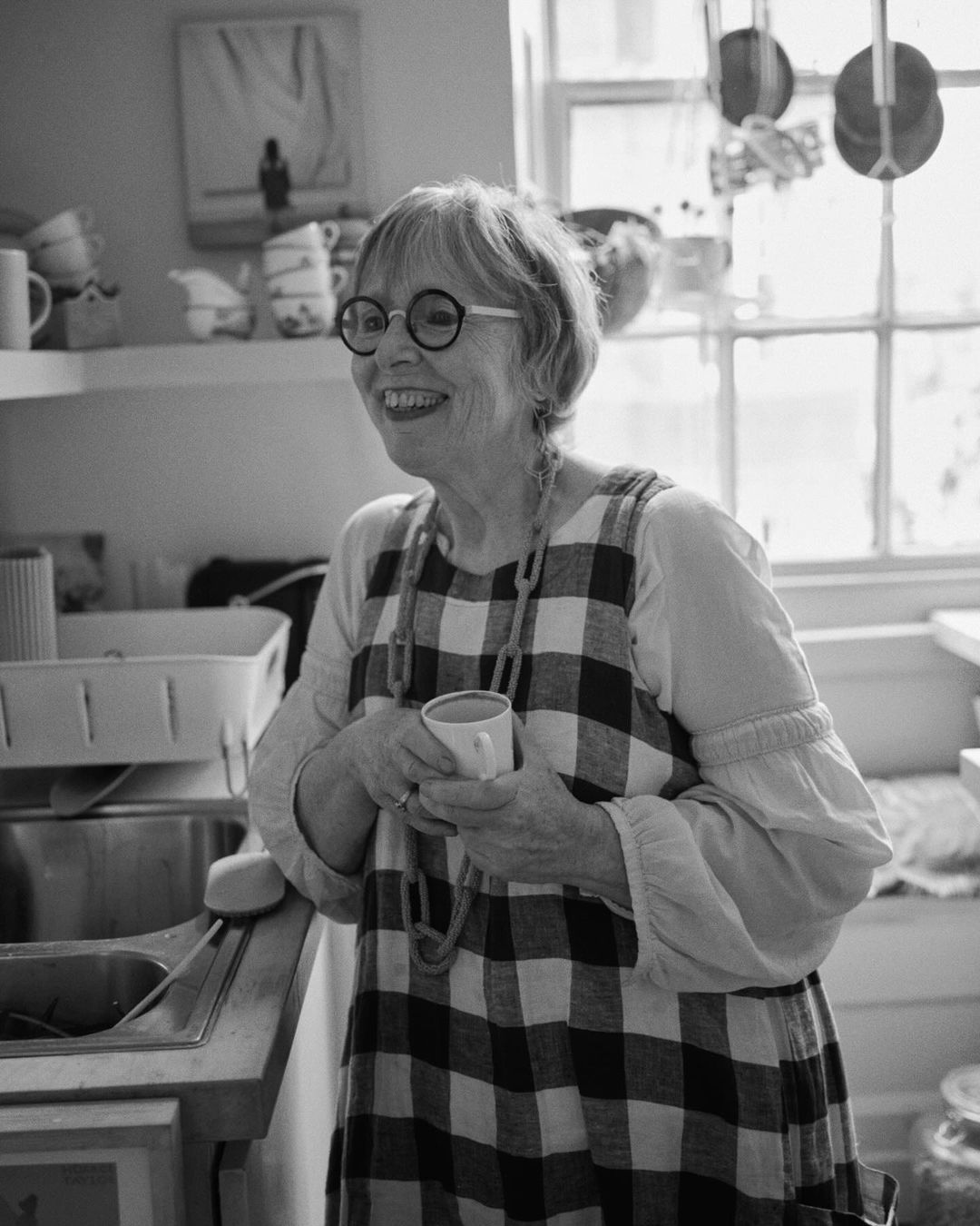 Older woman smiles in a bright kitchen, wearing glasses and a checkered apron, holding a mug near a sink and window.