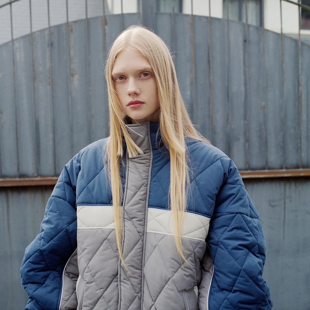 Portrait of a fair-skinned woman with long blonde hair wearing a quilted color-block jacket against a metal fence background.