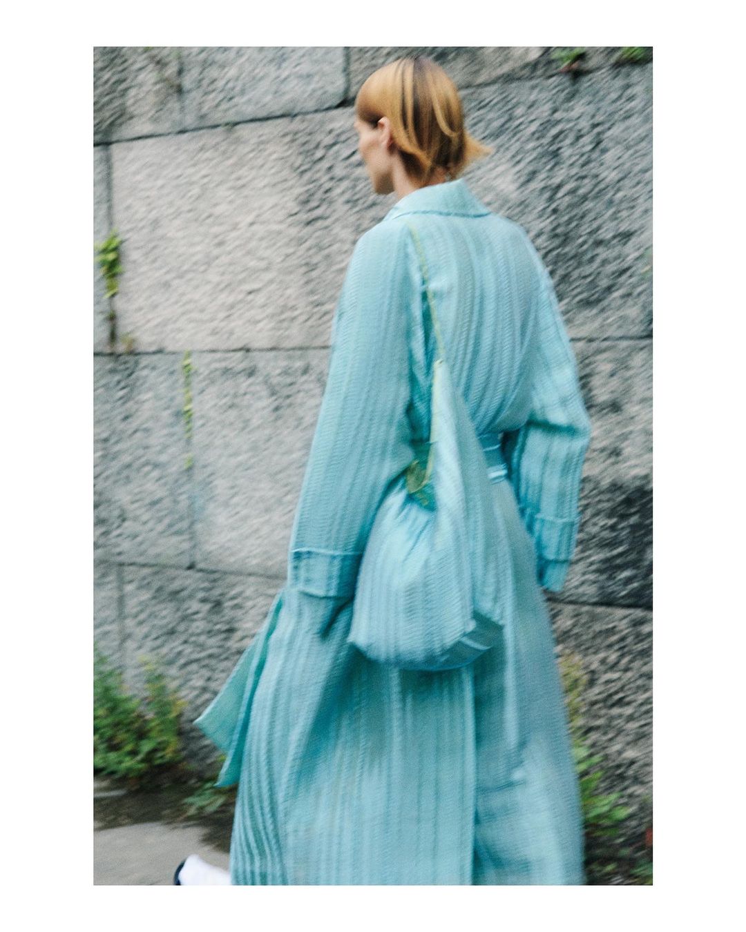 Woman in a light blue outfit walks away from camera carrying a matching purse against a textured concrete wall outdoors.