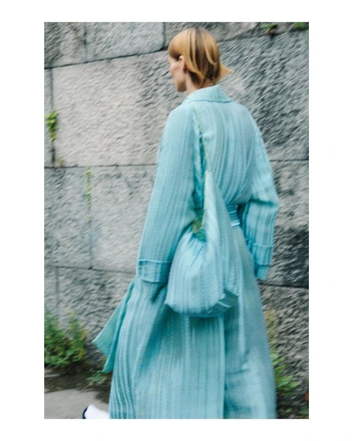 Woman in a light blue outfit walks away from camera carrying a matching purse against a textured concrete wall outdoors.