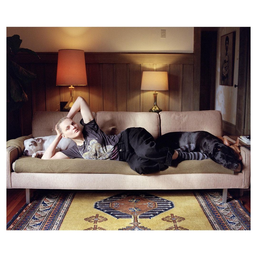 Woman lies on a couch with two dogs in an indoor setting featuring warm lighting, patterned rug, and wood-paneled walls. Relaxation concept.