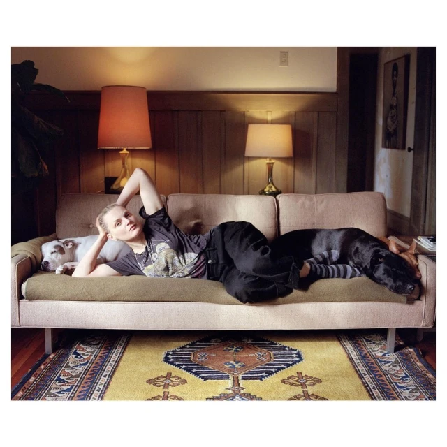 Woman lies on a couch with two dogs in an indoor setting featuring warm lighting, patterned rug, and wood-paneled walls. Relaxation concept.