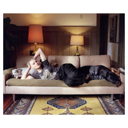 Woman lies on a couch with two dogs in an indoor setting featuring warm lighting, patterned rug, and wood-paneled walls. Relaxation concept.