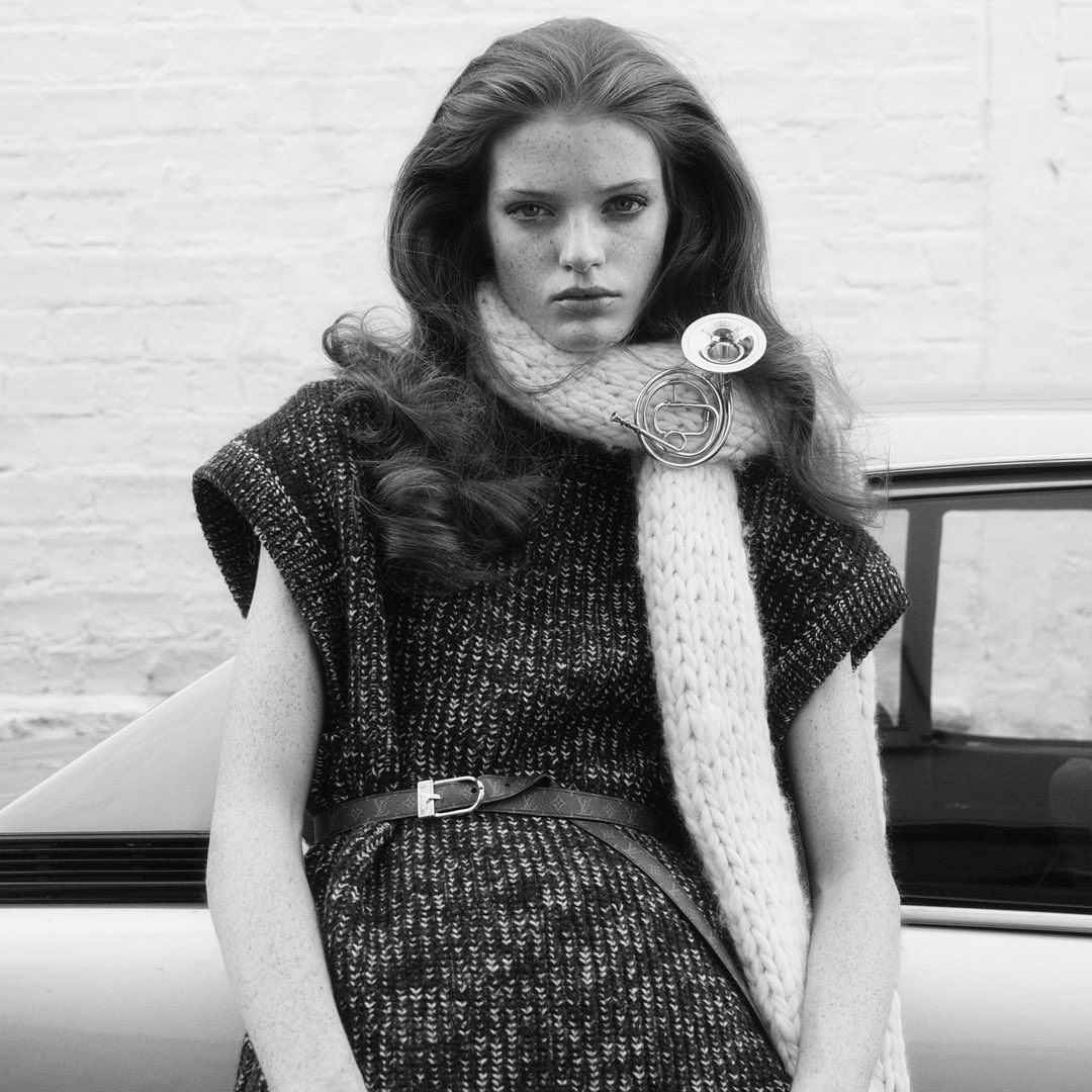 Model with long wavy hair wearing a knitted scarf and patterned dress leaning against a vintage car