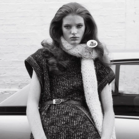 Model with long wavy hair wearing a knitted scarf and patterned dress leaning against a vintage car