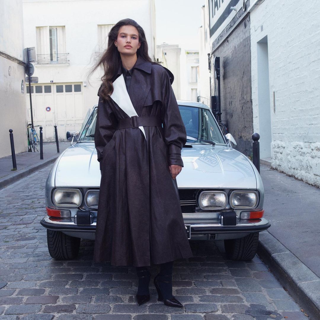 Fashion editorial of a woman posing in a brown trench coat and boots in front of a silver car on a European city street.