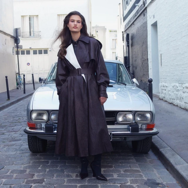 Fashion editorial of a woman posing in a brown trench coat and boots in front of a silver car on a European city street.