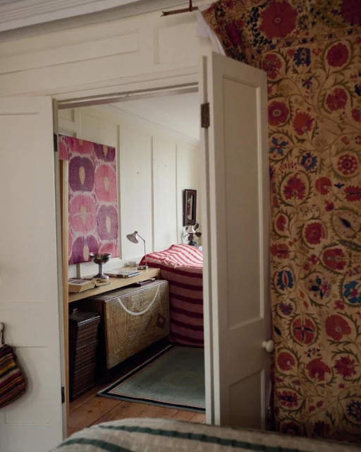 View of an interior room through a doorway with a bed, desk, and patterned textiles hanging on the wall creating a cozy and colourful space.