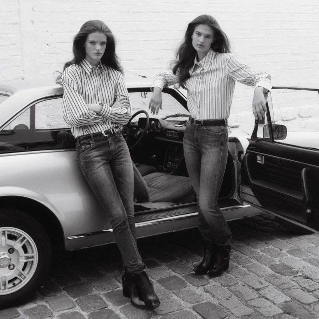Two women pose by a vintage car on a cobblestone street. They wear jeans, striped shirts, and boots in a black-and-white editorial shot.